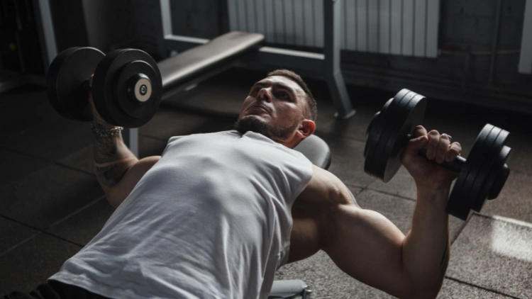 A man wearing white tank top is lying down doing a dumbbell chest press in a gym with other equipment in the background.