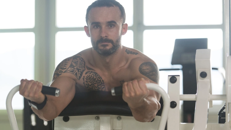 A topless muscled man with beard and tattoo is performing an exercise on a preacher curl machine in a gym with windows at the background.