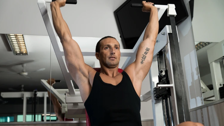 A muscled man with tattoo on his arm wearing a printed black tank top, performing a seated overhead shoulder press using the machine in a gym.