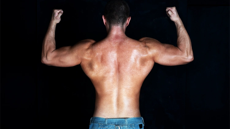 A back view of a topless man wearing a denim pants flexing his muscles with black background.