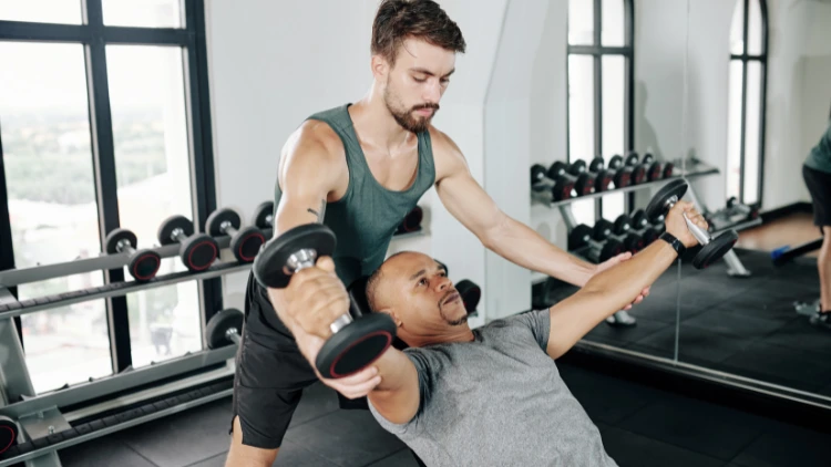 Man in green tank top with beard is healping the man in gray shrit to perform the dumbbell fly exercise at the gym.