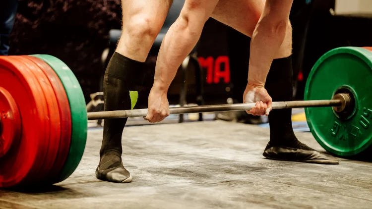 A man wearing a deadlifting shoes and shin guards in a deadlifting competition as he prepares to lift a heavy weight barbell with four weighted plates on each side, with colors green and red.