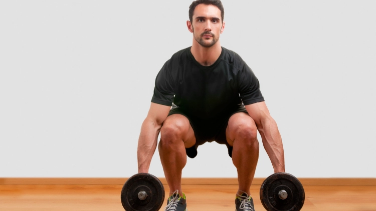 A man in black shirt and black short standing while in squat position holding dumbbell with both hands with a plain white background.