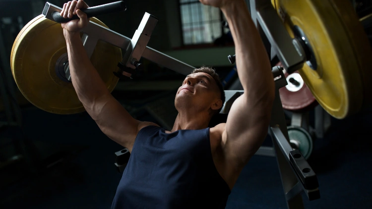 Man in blue tank top is doing a Iso lateral chest press with color yellow plate on the machine in a dimly lit environment.