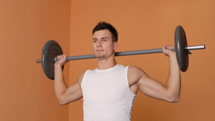 Man in white tank top is performing a behind neck press exercise in a brown background.