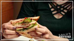 A woman wearing a green top is eating a McAlister's Keto entrée at a table with a holding a club sandwich, the sandwich has ham, cucumber, lettuce and slightly toast bread, rings and nail colors are visible from the woman's hand.
