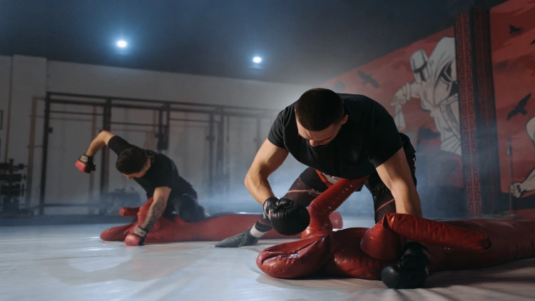 Two men wearing black shirts and fitted gym tights are shown throwing punches at human dummies placed on the floor in a gym with monkey bars visible in the background.