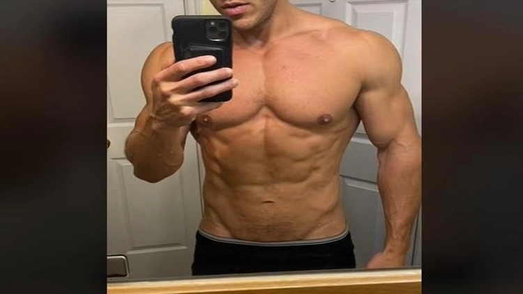 A man taking a picture of himself using his phone with a black case in the mirror, flexing his muscles without a shirt on.