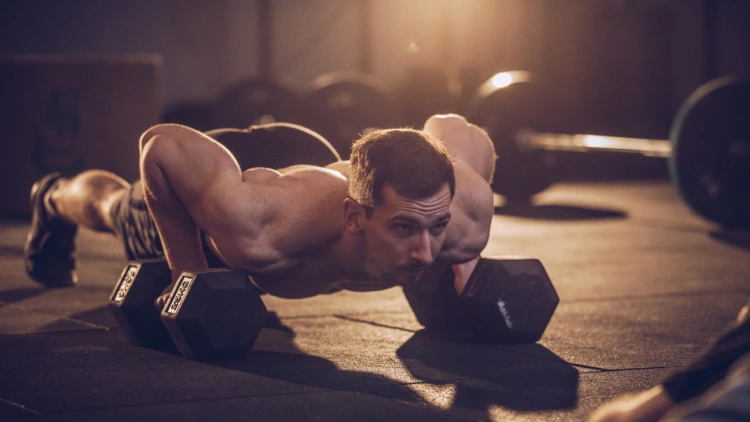 A muscular man is performing dumbbell push-ups, he is in a plank position with each hand holding a dumbbell, placed on the ground slightly wider than shoulder-width apart, he is lowering his chest towards the ground, bending his elbows and keeping his body straight. 