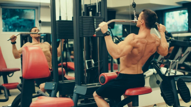Topless guy in black shorts doing a lat pulldown exercise at the gym with mirror on his front and other gym equipment are seen in the background.