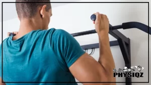 A black man is using his biceps and lats, two of the neutral grip pull up muscles worked, to pull his chin above a metal bar and he's wearing a dark cyan t-shirt and facing a white wall.