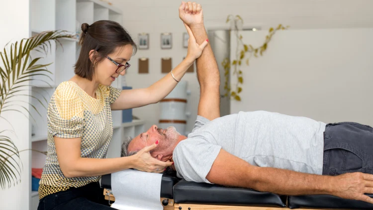 An elderly man in a white shirt lies on his back on a medical examination table, a therapist in polka dots top sits beside him, checking his wrist with one hand while the man raises his other hand towards the ceiling.