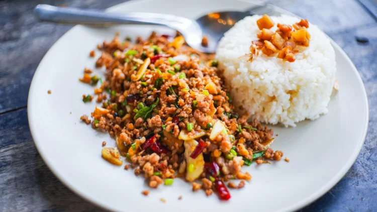 A white plate with textured edges is filled with a Thai basil pork dish made with pork, fresh herbs, and vegetables, accompanied by a side of fluffy white rice.