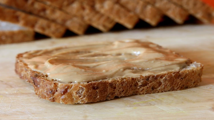 A slice of wheat bread with a layer of peanut butter positioned on a wooden surface, with additional slices of wheat bread visible in the background.
