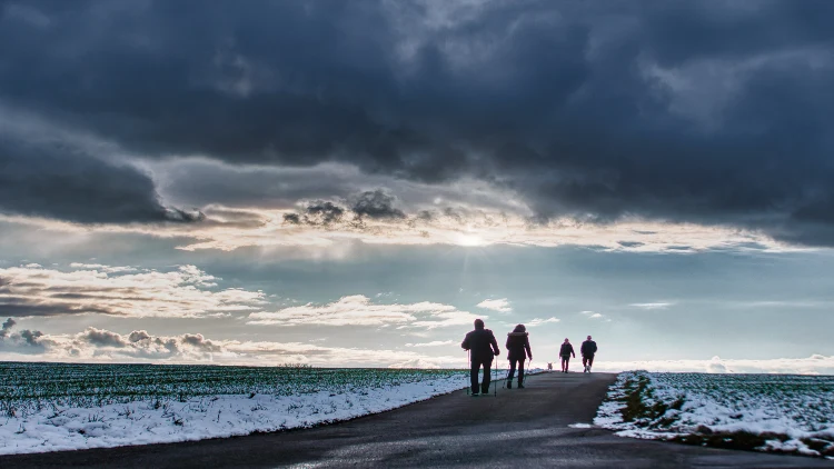 Four people wearing winter attire are walking down a snowy road, with clouds in the sky above them.