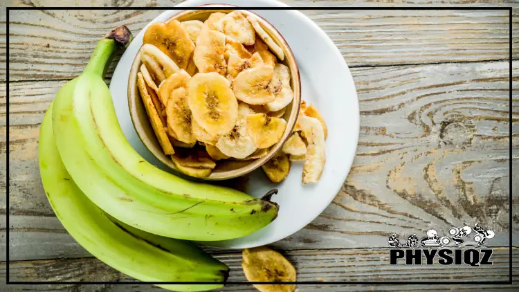 A brown bowl filled with sliced, dehydrated plantains sits on a white plate next to two fresh green plantains, all displayed on a wooden surface.