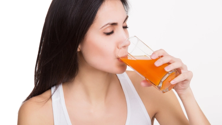 A pretty woman wearing a white tank top is drinking an orange-colored drink from a clear glass, with white background.