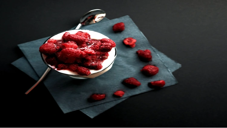 A serving of yogurt topped with fresh raspberries, presented on a gray napkin with a silver spoon, all set against a plain black background.