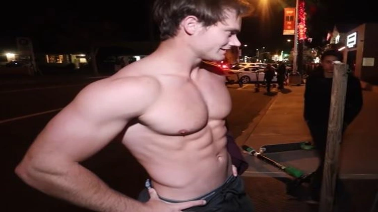 A man stands shirtless on the street with his abs flexed, displaying well-defined muscles.