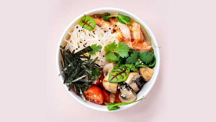 A white bowl containing angel-hair pasta noodles, shrimps, diced mushrooms, baby tomatoes, seaweeds, and fresh parsley, garnished with black sesame seeds, is set against a white background.