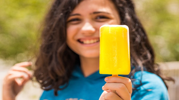 A young girl with short hair, wearing a blue shirt and smiling while holding a yellow ice popsicle appears to be enjoying the treat, with the background blurred to make her and the popsicle the focal point of the image.