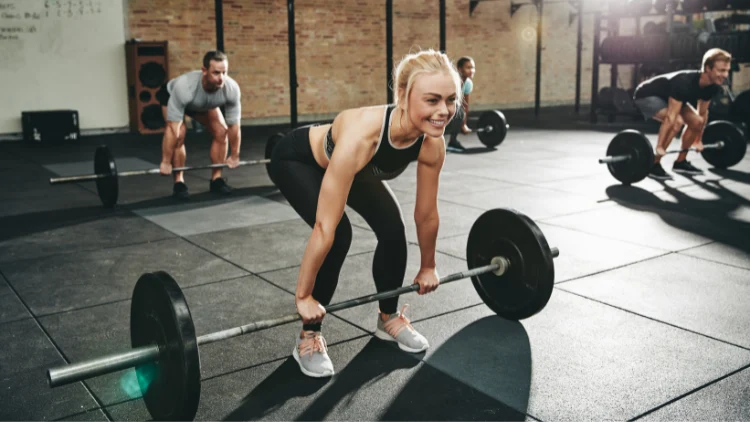 A woman smiling wearing a black top, black pants, and gray shoes performing the power clean in starting position, with the barbell on the floor, standing with feet hip width apart, bending at the knees and hips and grabbing the bar in a hook grip, in a gym.