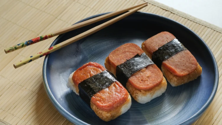 Three servings of spam musubi on a black plate with chopsticks on a wooden placemat.