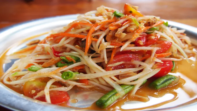 A spicy green papaya salad with unripe papayas, chili pepper, fish sauce, lime, palm sugar, garlic, and raw Thai eggplant served on a silver plate on a wooden table.