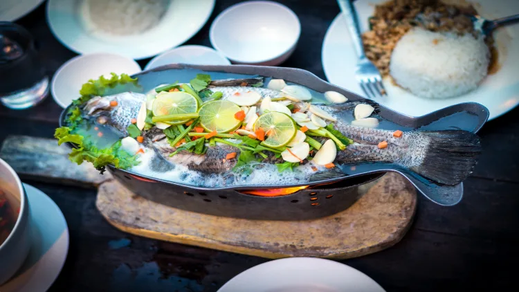 A platter of steamed fish with a zesty lime and garlic sauce heated on a charcoal grill, served on a wooden board with small bowls and plates in the background.