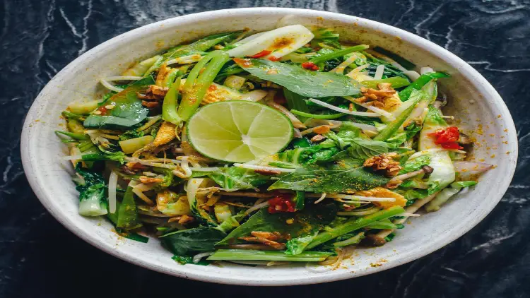 A serving of a Thai beef bowl made with a pound of ground beef mixed with fresh basil, red pepper flakes, and vegetables such as finely chopped carrots, scallions, bean sprouts, limes, cucumbers and spinach, served on a white bowl atop a black marble surface.