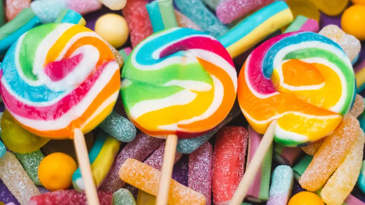 Three round candies in rainbow colors, attached to sticks and on top of a pile of small, sugar-coated candies.