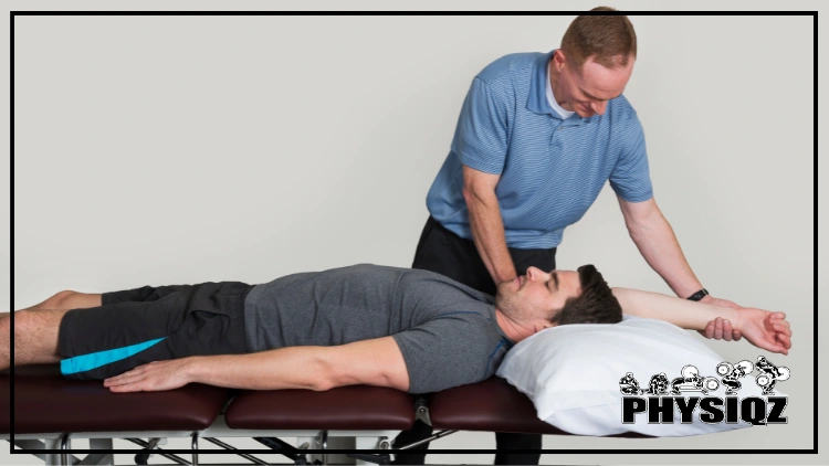 A therapist in a blue shirt is assessing a man lying on his back in a gray shirt and black shorts, with a white pillow under his head; the therapist is holding the man's extended arm, checking for tightness in his lat muscles.