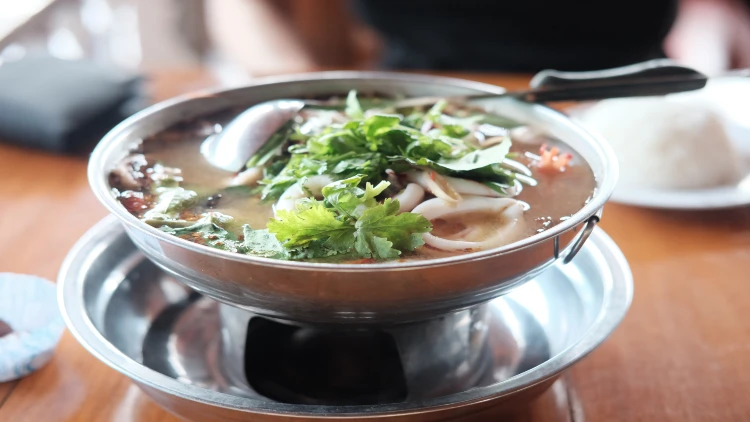 A bowl of soup with fresh lemongrass, fish sauce, chilies, kaffir lime leaves, and shrimp, served with a silver bowl underneath and put on a table.