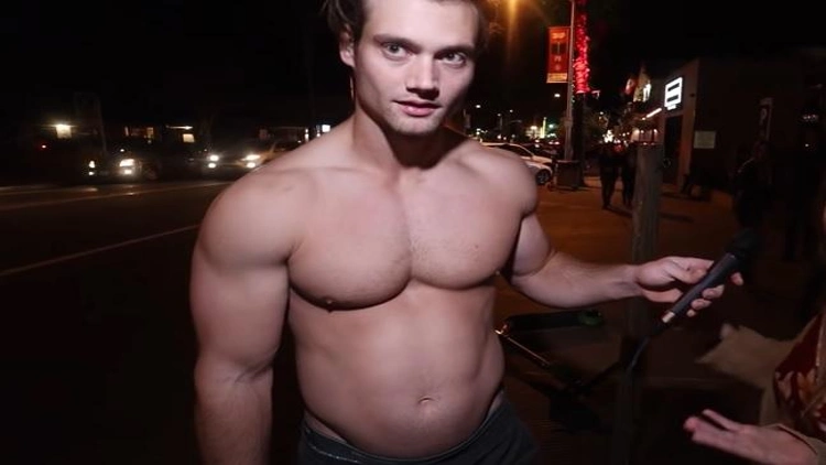 A shirtless man with muscular physique standing on the street holding a microphone.