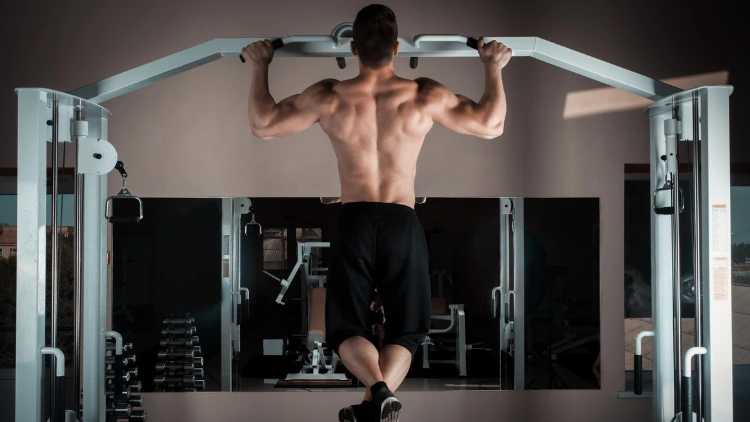 A back view of a topless man wearing a black pants and shoes performing a wide grip pull up exercise using a pull up machine in a gym.