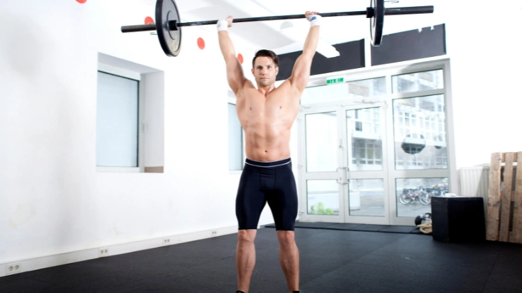 Topless man in black shorts doing push press with white wall and big glass door in the background .