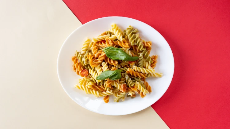 A white plate with tri-color rotini pasta topped with fresh basil, set against a background of white and red colors.