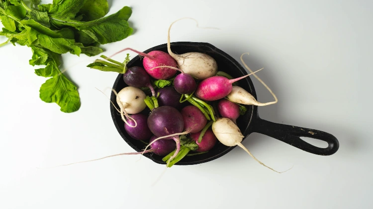A black frying pan holds a colorful assortment of freshly harvested turnips in shades of purple, pink, and white, with vibrant green turnip leaves visible in the white background.