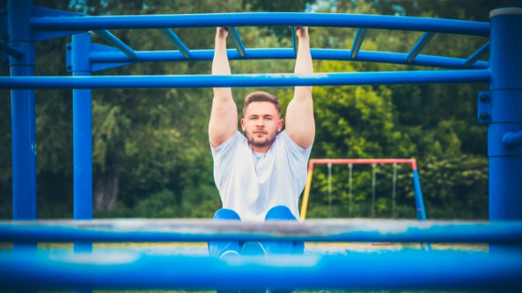 A man wearing a white shirt and blue pants is shown doing close grip pull ups on a blue metal bar at a park, the background consists of green trees and grass, while the focus is on the man's upper body strength as he pulls himself up towards the bar.
