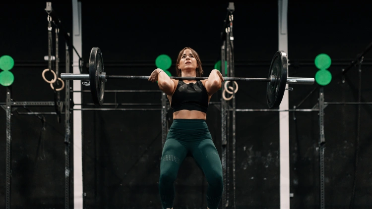 A woman wearing a black top, green pants is performing the hang clean in second position, with her arms guiding the barbell in a gym.