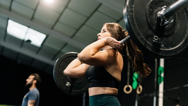 A woman wearing a black top, green pants, performing the power clean in final position, standing up straight into full knee and hip extension with the bar in rack position, completing one rep, in a gym.