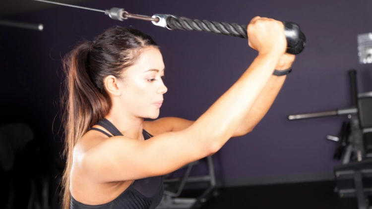 A woman working out at the gym, performing an overhead cable extension exercise, she is using a cable machine with the handle held above her head, while extending her arms upwards, her toned arms is visible.