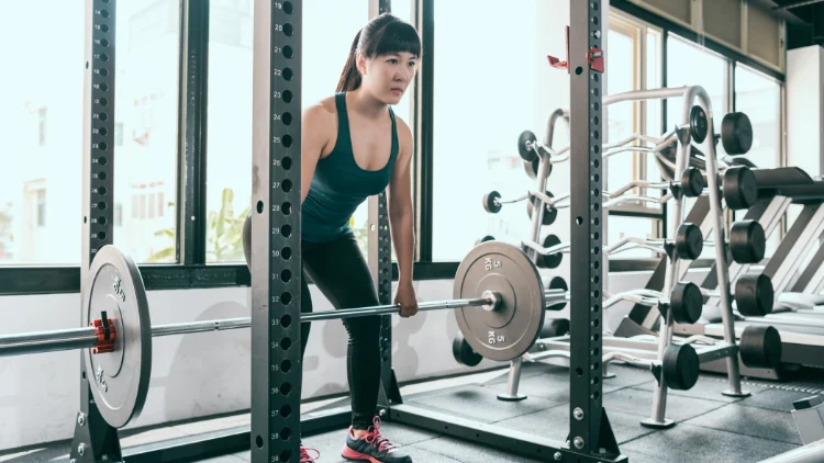 A asian woman wearing black tank top and black pants performing rack pulls exercise with a barbell in a gym with various gym equipment in the background.