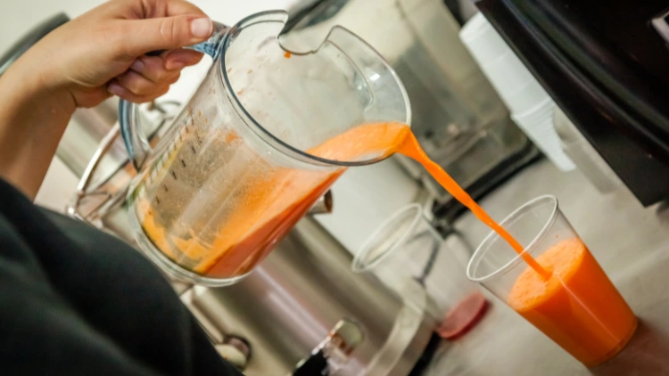 Staff member in a pouring a orange smoothie from a blender into a clear plastic cup, the smoothie appears to be a thick and creamy consistency with a bright orange color.
