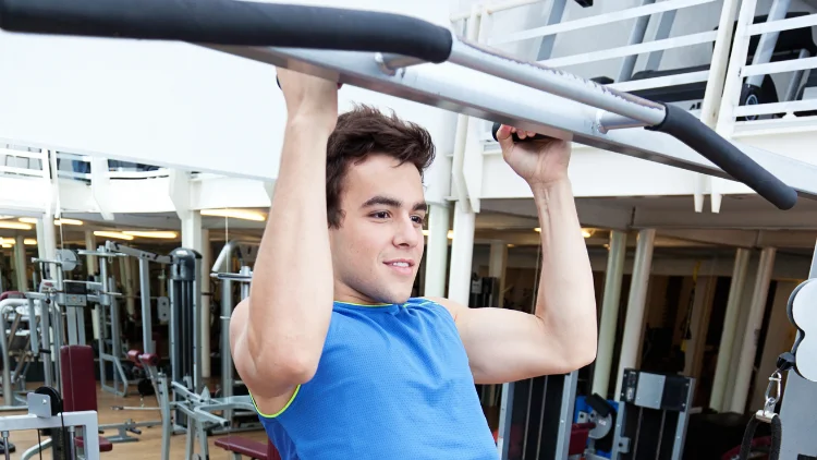 A young man wearing a blue tank top is performing pull ups exercise using a neutral grip at the gym.