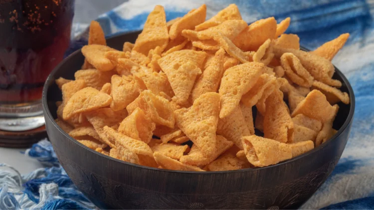 A black bowl filled with Bugles chips, with a refreshing glass of soda in the background, the golden-brown chips are arranged in a mound, with their signature cone shape and ridges visible, the carbonated drink in the glass creates a contrast to the savory and crunchy snack, providing a balance of flavors.