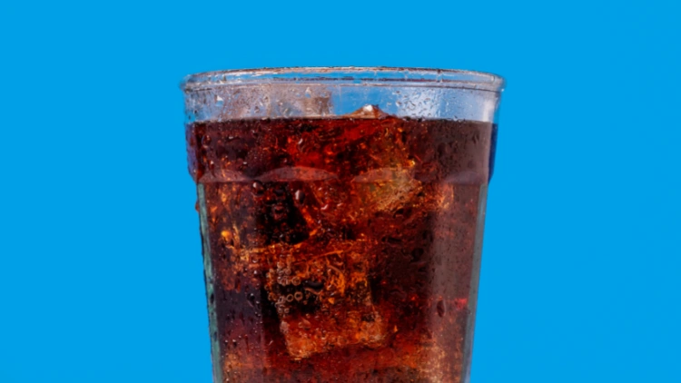 A moist, clear glass filled with cold carbonated soda and ice cubes displayed against a sky-blue backdrop.