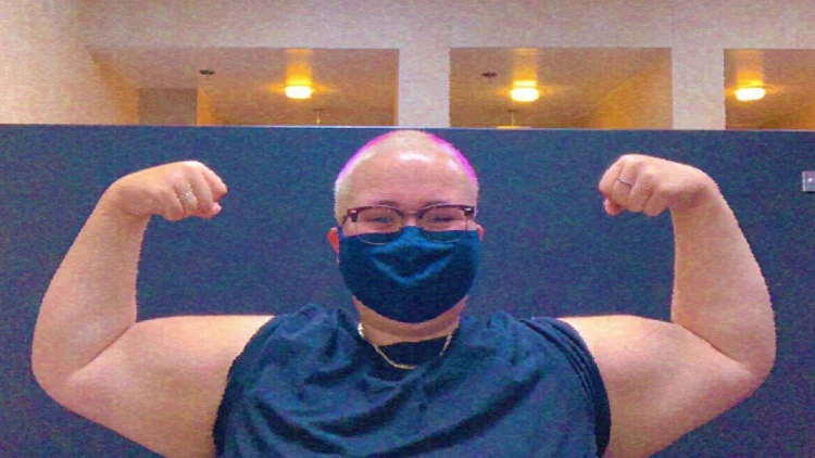 A man wearing a blue shirt and a blue face mask, flexing his arm with visible fat on it, the man's arm is visible in the foreground of the image, with some fat and little muscle definition.