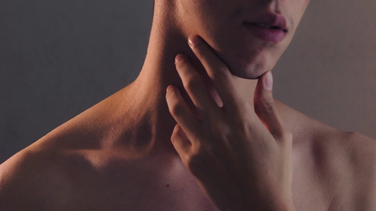 A close-up photo of a skinny topless man holding his chin in a dimly lit studio.