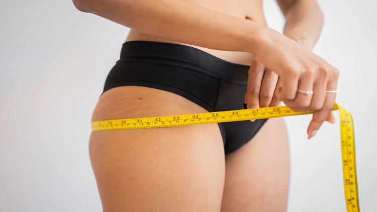 An image of a woman wearing black undergarments and having two rings on her hands is measuring her thighs using a tape measure against a white background.